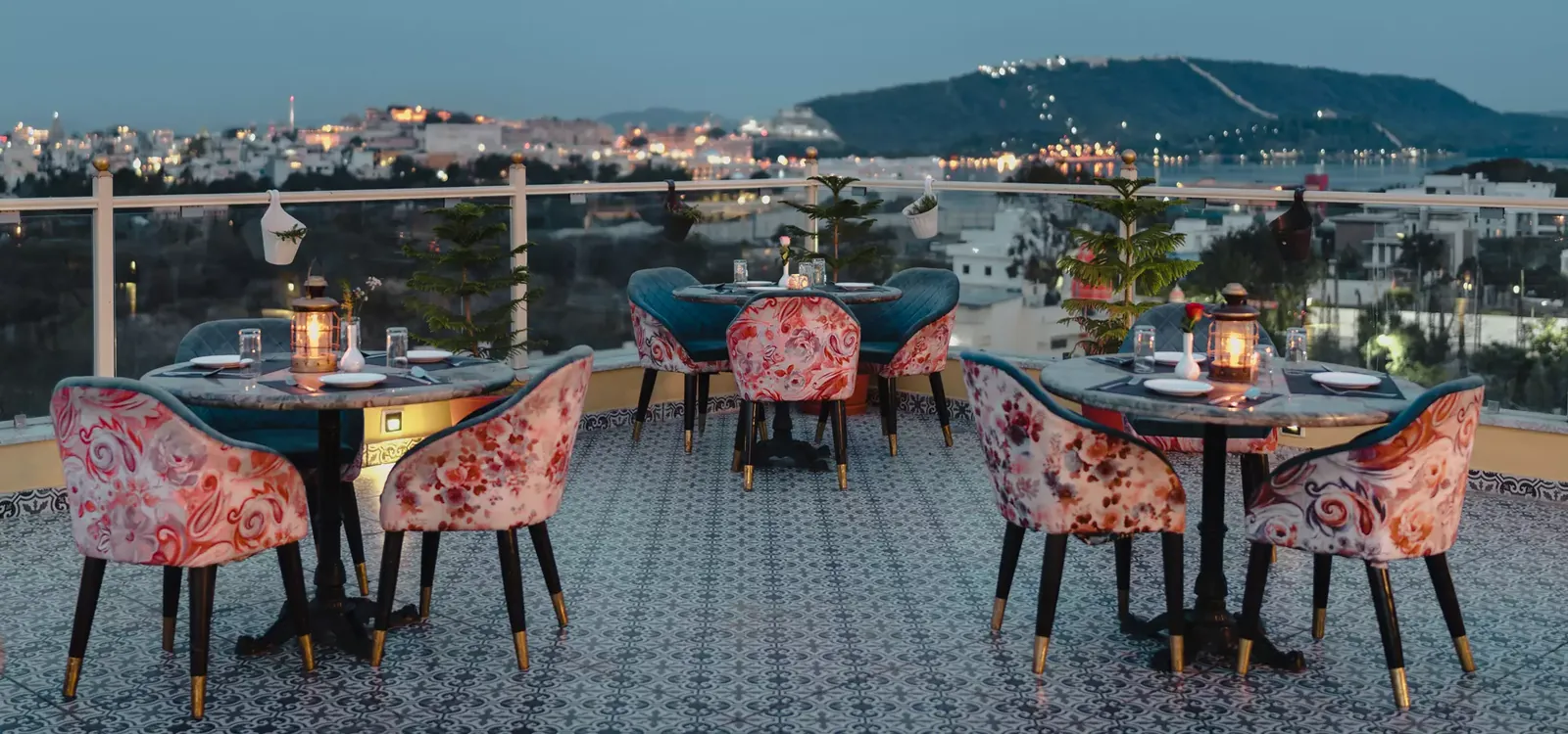 Rooftop tables at The Chronicles Hotel at dusk, with the lit City Palace and Aravalli hills across Udaipur